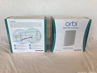 Two Netgear Orbi Satellite boxes side by side showing front and side views with branding and specs visible.