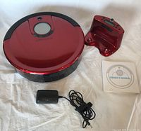 Red Bobsweep robotic vacuum, matching red docking station, owner's manual, and black power supply adapter laid out on a white surface.