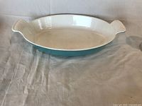 Oval Le Creuset casserole dish sitting on a textured cloth, showing interior cream-white enamel with some minor marks and original shape and handles.