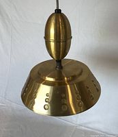 Close front view of the brass pendant light showing the flying saucer shaped perforated shade and bullet-shaped adjustable section.