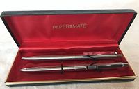 Pen and pencil set in open box showing brushed stainless steel finish and red interior lining with Paper Mate logo.