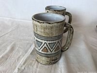 Two Mid-Century Modern ceramic mugs with textured glaze; one with zigzag pattern, one with oval leaf pattern, displayed from front angle.