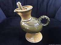 Side view of the pottery pitcher showing bulbous olive green body, pedestal base, and tentacle-inspired circular handle.