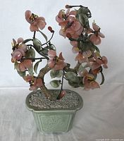 Full front view of the vintage art glass tree in a celadon green ceramic planter with pebble-like filler and pink flowers with green leaves.