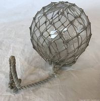 Clear spherical vintage glass fishing float enclosed in beige woven rope net with loop handle, lying on white fabric.