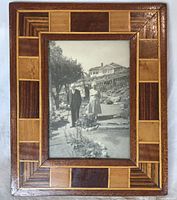 Frontal view of the rectangular marquetry frame showing wood inlay geometric pattern and centered vintage black and white photo of a man and woman.