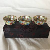 Three hand painted vintage Chinese porcelain cups with floral and bird motifs on a black silk fabric box with red patterns.