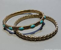 Two gold-tone metal bangles, one with turquoise-colored oval stones and the other with a geometric diamond pattern, shown stacked.