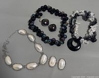Three costume jewelry necklaces and earrings displayed on gray surface: Silver-tone oval pearl-like stone necklace with matching earrings; black iridescent bead necklace with matching earrings; multi-strand black and clear chip bead necklace with black circular pendant.
