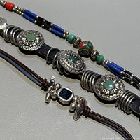 Close-up showing three bracelets; silver bracelet with turquoise stones, brown leather bracelet with dark stone, beaded bracelet with blue and turquoise beads.