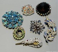 Seven vintage rhinestone brooches assorted styles and colors laid out on grey background.