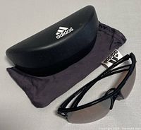 Black Adidas sunglasses with brown lenses, hard case, and soft pouch displayed on gray surface.