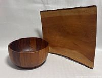 Serving bowl in darker teak wood with polished finish next to the live edge wooden tray with natural bark edge and smooth light wood finish.