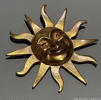 Gold-tone sun-shaped brooch with smiling face shown against a neutral background. The brooch features detailed facial features and stylized sun rays with alternating curved and pointed designs.
