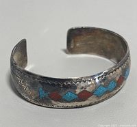 Close-up front angle of sterling silver open cuff bracelet with red and turquoise diamond inlay pattern and engraved zigzag motif on the sides.