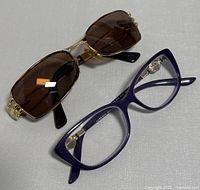 Pair of Versace eyeglasses with purple frames and vintage Sover sunglasses with gold frames and brown lenses placed side by side on a gray surface.