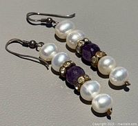 Close-up image of earrings showing white pearls, faceted purple amethyst center beads, and rhinestone spacer rings on silver hooks.