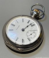 Front and side angle showing white dial with Roman numerals, small seconds dial, and silver case with crown and loop.