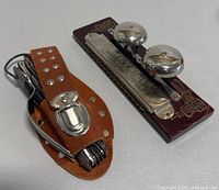 Foldable metal camping multi-tool with utensils secured in a brown leather case, and a silver harmonica with two bells on a wooden base seen from a side angle.