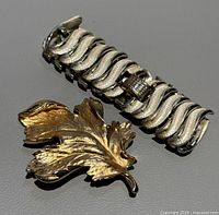 Silver-tone wavy patterned bracelet and gold-tone leaf-shaped brooch lying on gray background showing condition and design details.