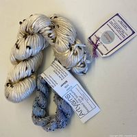 Two skeins of silk yarn, one cream with brass sequins, one pale grey with silver sequins, with original tags attached.