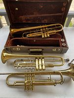 Overview showing three brass trumpets on table and trumpet in case, plus two mouthpieces.