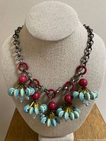 Front view of necklace on display bust, showing five clusters of light blue ceramic stones with red beads, hanging from a metal chain with red rings.