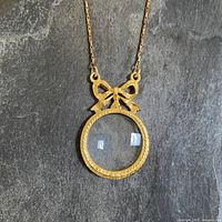 Close-up front view of the gold tone magnifying glass pendant with bow, showing the texture of the metal and clarity of the lens.