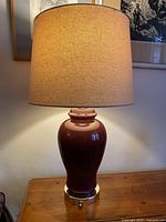 Full view of the lamp showing the dark cherry ceramic body, brass base, and new beige shade on a wooden table.