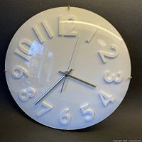 Front view of the round white wall clock showing raised numbers and clear convex cover with silver hands.