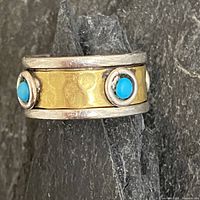 Close-up of sterling silver and brass spinner ring with turquoise stones on a dark stone background showing front and side views of the spinner.