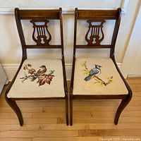 Pair of wooden chairs with carved backs and bird tapestries on seats placed side by side on hardwood floor.