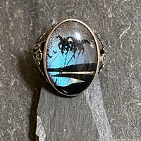 Front view of oval ring showcasing the iridescent butterfly wing with a tropical scene under concave glass and detailed silver floral sides.