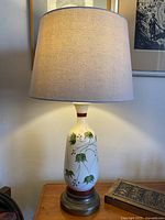 Full view of the vintage hand painted ceramic lamp with beige fabric shade, showing lamp lit and ambiance on wooden table