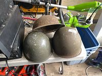Three helmets on a table: Brodie helmet showing rust, two M1 style helmets olive drab