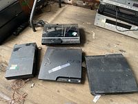 Four Sony PlayStation consoles arranged on a wood floor: PS2 fat (vertical), PS3 fat (horizontal), PS3 Slim, PS3 Super Slim