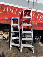 Both aluminum step ladders standing side by side against a truck for size reference