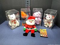 Wide shot of all six items: four Ty Beanie Babies in clear plastic display cases, one Santa plush, and an unopened pack of Official Ty Collectors Cards.