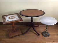 Three plant stands grouped together: square ceramic tile top, oval inlaid wood top, round marble top with brass base