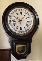 Full view of the antique E. Ingraham Dew Drop calendar wall clock mounted on the wall showing round face with Roman numerals and calendar markings, and lower pendulum window.