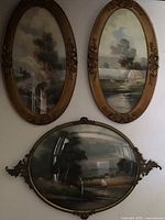 Photo showing the trio of framed oval pastel paintings on a wall: two long matching gold painted frames on top, one larger decorative gold metal framed oval painting below