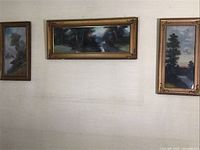 Three antique pastel landscape paintings hung on a wall, arranged with two vertical paintings flanking a larger horizontal painting in matching gold leaf frames with metal corner ornaments.