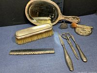 Complete antique sterling silver vanity set on blue fabric including hand mirror, brushes, grooming tools, and perfume bottle.