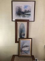Three framed winter scene pastel paintings stacked against a wall, showing size and frame details.