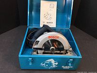 Bosch circular saw inside blue metal carrying case with instruction manual on lid.