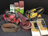 Overview photo showing the DeWalt orbital sander, Skil belt sander, assorted sandpaper belts, discs, and sanding pads arranged on a surface.