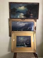 Three framed antique pastel ocean scene paintings stacked vertically on a wall. Top painting with a dark wooden frame depicting a full moon and a figure in a hut. Middle painting with a wide gold frame and ornate corners shows cliffs and ocean waves. Bottom painting with a gold frame and metal corner decorations shows a moonlit ocean scene with crashing waves.