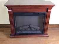 Front view of electric fireplace in wood case showing carved side columns and glass front with faux logs inside.