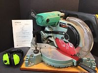 Hitachi Koki C10FCD 10-inch compound table saw with red blade guard and clear plastic cover, alongside instruction manual and radio headphones.