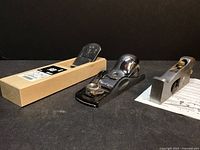 Three woodworking planes including a Japanese wooden block plane, a Woodriver metal plane, and a Stanley #92 shoulder plane arranged on a black surface.